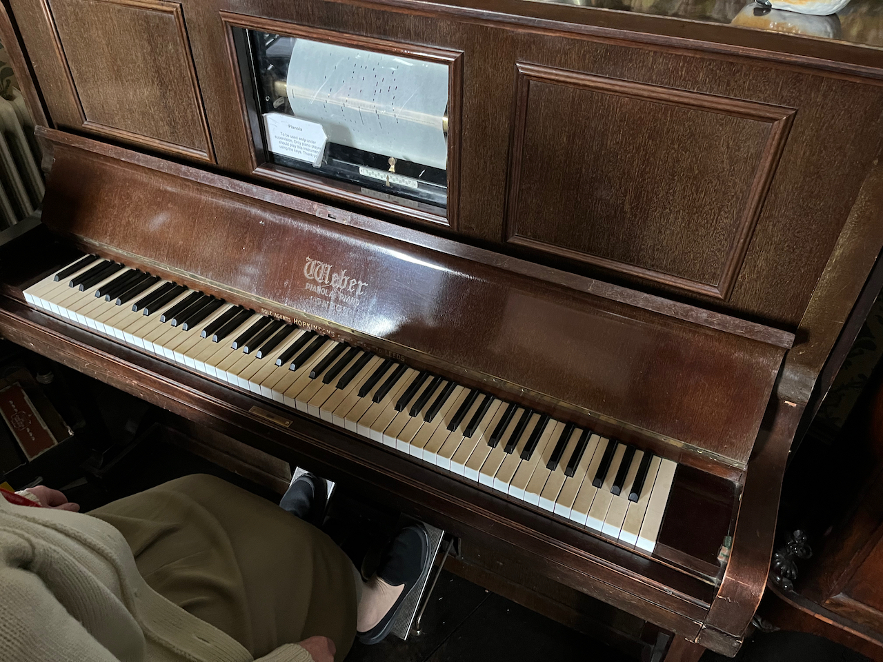 A pianola. This is a large wooden piano with pedals underneath. When the pedals are pressed, a roll of card with punched holes rotates in the window visible on the top panel of the pianola, and the keys below play themselves.