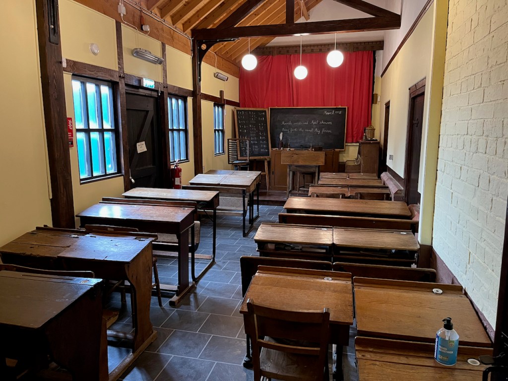 An old school room, with rows of wooden desks and chairs facing 2 blackboards at the far end, one on a pedestal displaying some maths, and another on the wall showing a bit of poetry, that says March winds and April showers, bring forth the sweet May flowers.
