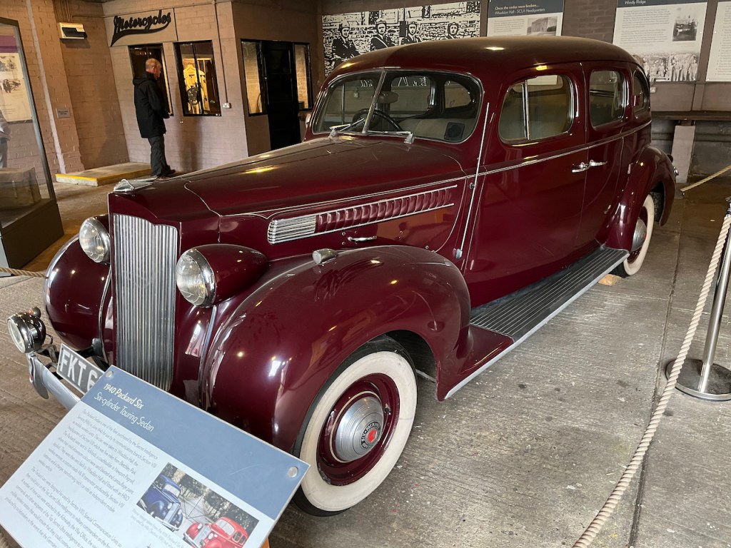 A 1940 Packard Six Sedan, a large red car with a tall metal grille at the front of its long bonnet, a large footplate by the doors along the side, and white rims on the tyres.