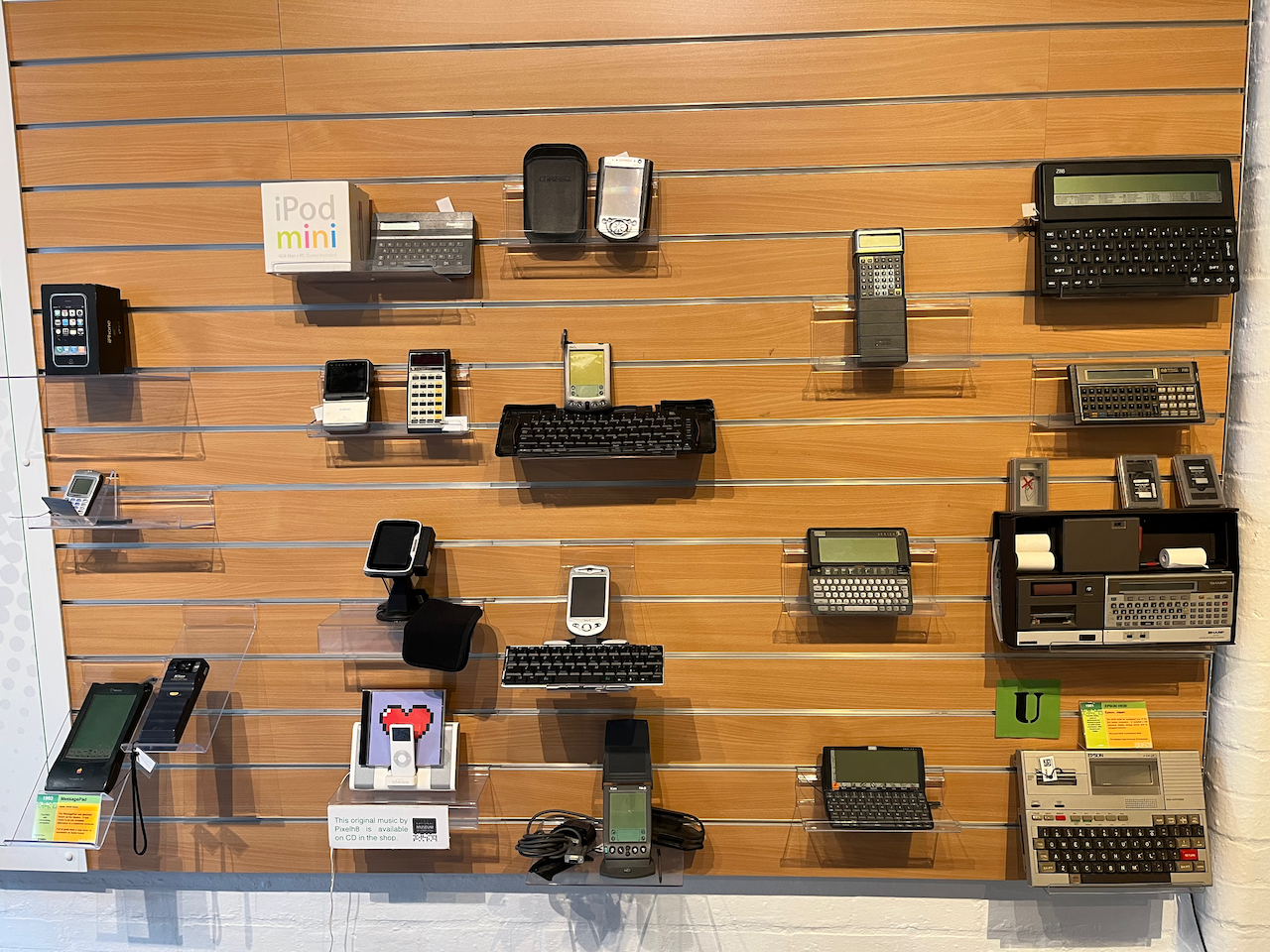 Wall display of early pocket computers and personal digital assistants, including early iPods, iPhones and a Message Pad by Apple, plus devices by Sharp, Hewlett Packard, Epson and others.