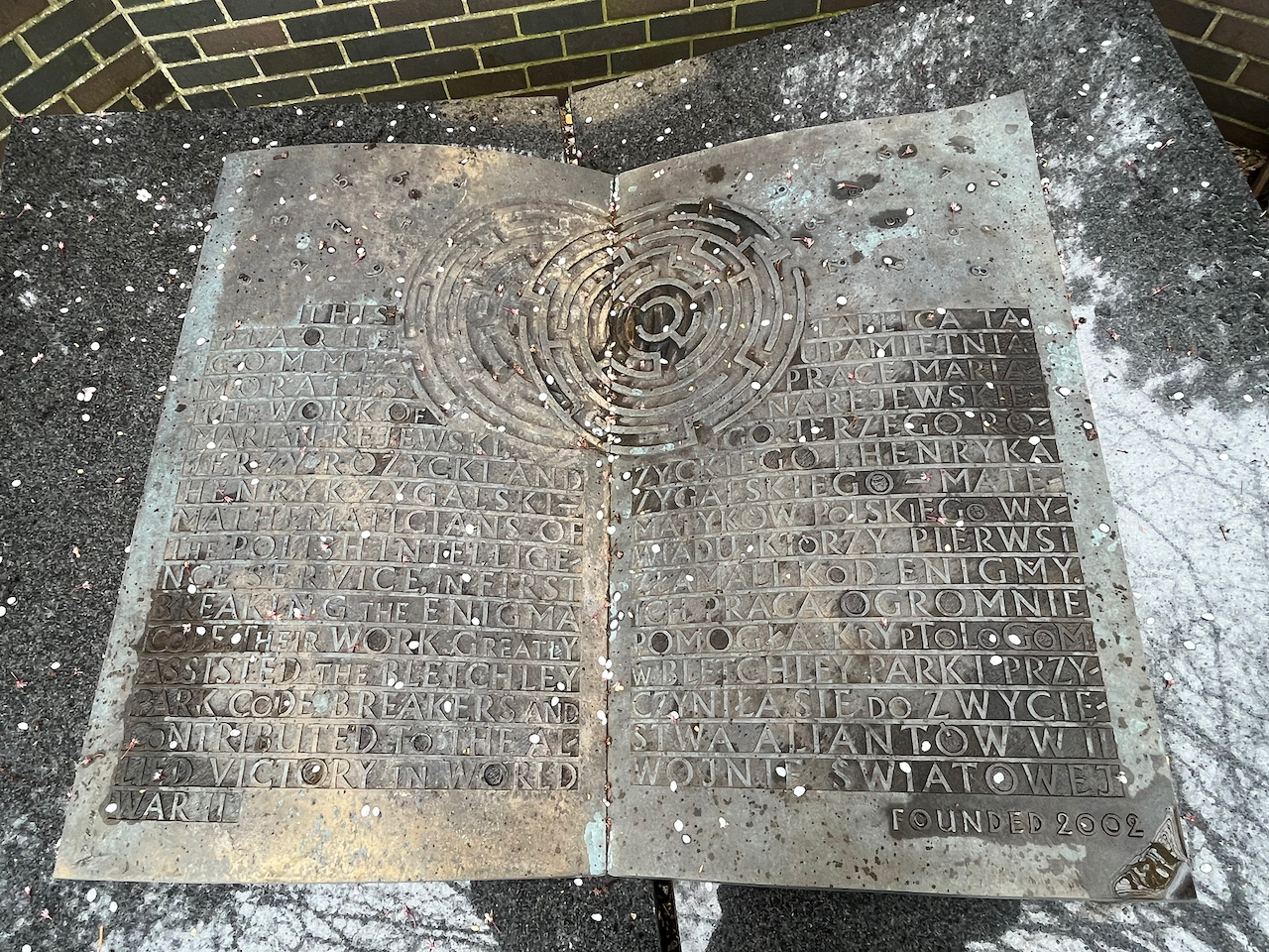 Large metal sculpture of an open book, with English writing on the left page, and Polish writing on the right, all in capitals. In the centre at the top is a circular maze design, spanning both pages so the text has to wrap around the sides of it.