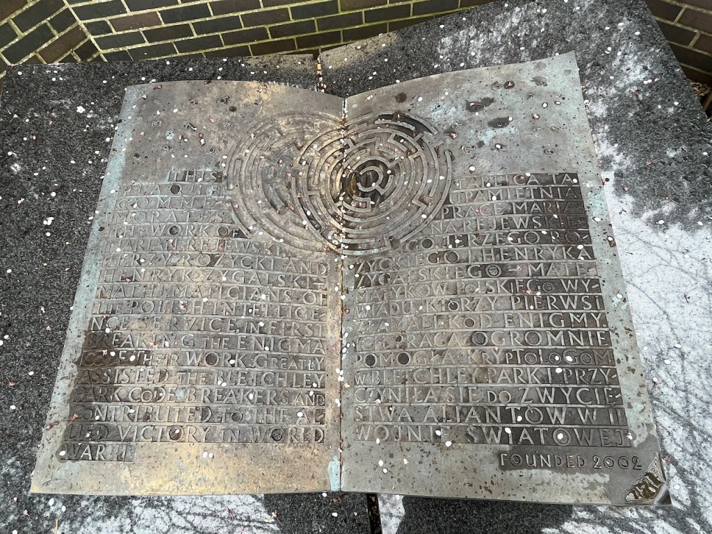 Large metal sculpture of an open book, with English writing on the left page, and Polish writing on the right, all in capitals. In the centre at the top is a circular maze design, spanning both pages so the text has to wrap around the sides of it.
