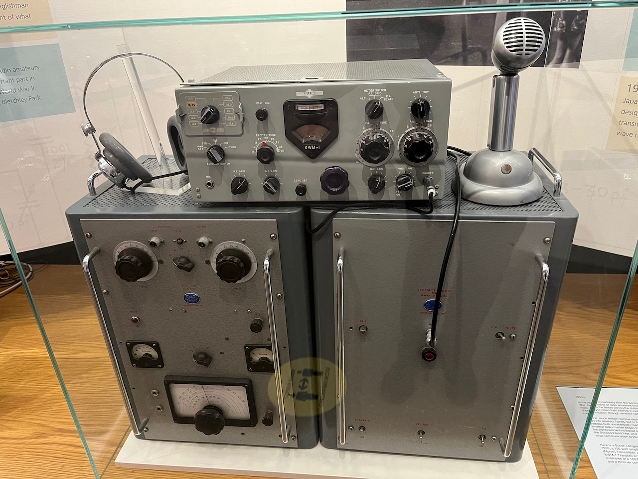 Radio equipment, consisting of 2 large metal boxes, the left of which has a few controls and a frequency display. On top of those boxes is a smaller metal box with lots of dials, plus a microphone and a pair of headphones.