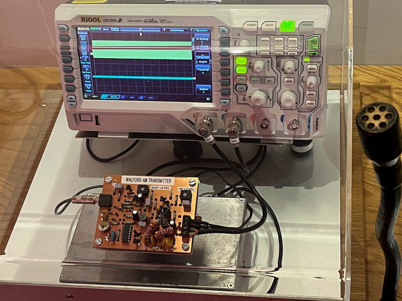 An oscilloscope unit, consisting of a box with a screen on the left, and lots of dials and buttons on the right. A small transmitter board with lots of components on it is sitting in front of the oscilloscope, connected to it by a cable.