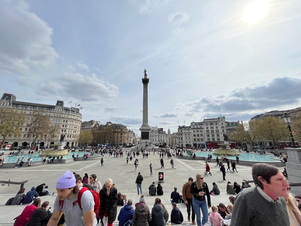 Trafalgar Square, a huge paved area with 2 large pools with fountains on each side, a large open paved area in the centre, and Nelson's Column at the far end by a large, busy junction. There are quite a few people, but it's not crowded.