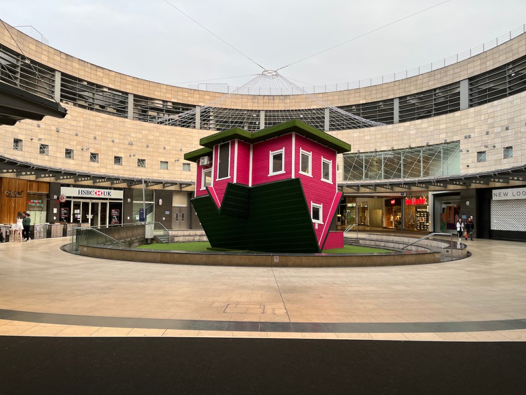 A large pink upside down house, with white edging for the walls, windows and doors, and a black roof. It's stood on a green surface in a large circular area, in the Midsummer Place shopping centre, surrounded by a wide pathway with shops.