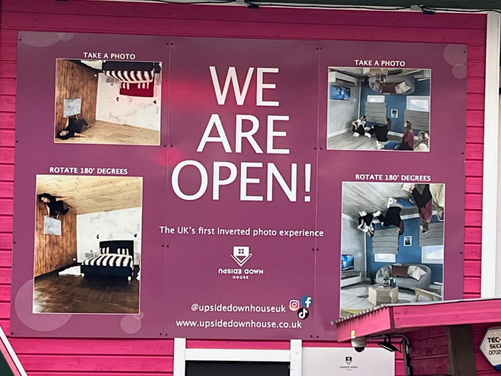 Close up of the sign above the entrance of the Upside Down House, saying We Are Open in white against a dark pink background. To the left and right of that are photo examples taken in different rooms in the house. In each case, the top photo shows people posing in the upside down room, and below that the same photo is flipped 180 degrees so it looks like they're hanging from the ceiling.