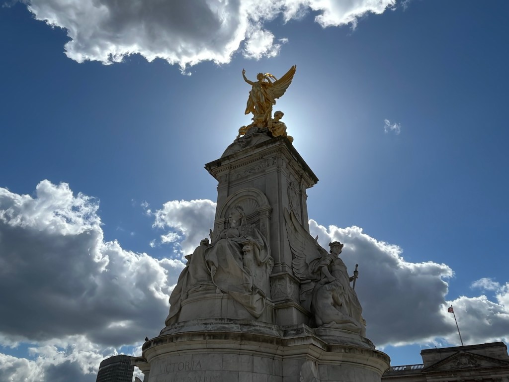The Queen Victoria Memorial, a huge stone column with statues around its wide base, including one of Queen Victoria herself, and a gold statue of a winged goddess on the very top of the structure, one arm held aloft.
