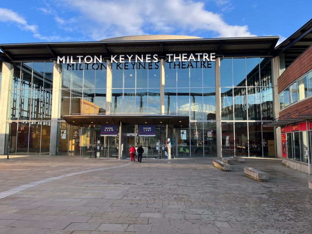 The exterior of the Milton Keynes Theatre, a large glass-fronted building, beneath a sunny blue sky. The name of the theatre is in large white capital letters across the top of the highest windows.