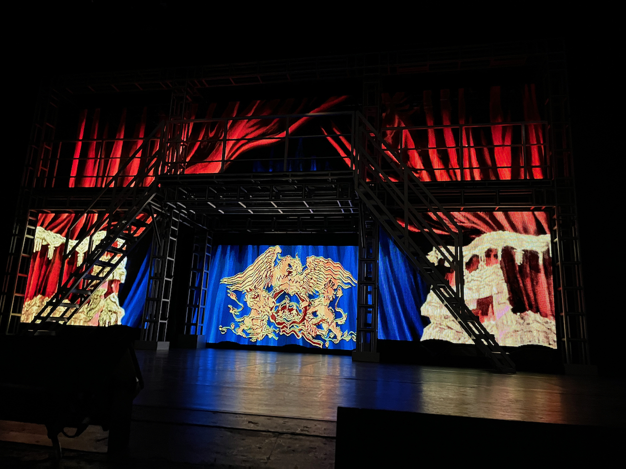 View of the stage at the start of We Will Rock You, with the ornate Queen crest lit up on a blue curtain at the back of the stage.