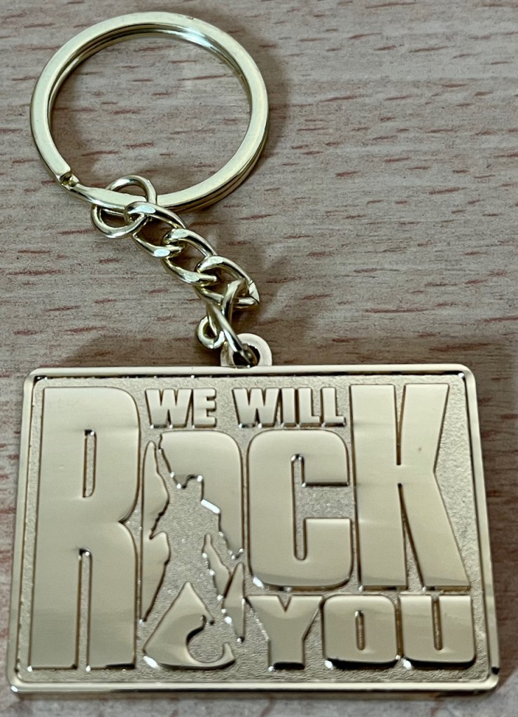 A gold keyring with the rectangular logo of the We Will Rock You Musical. The letter O in Rock, the largest word in the logo, has a silhouette of Freddie Mercury in the centre of it, standing legs apart with one arm punching the air triumphantly.