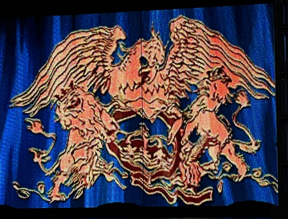 Close-up of the Queen crest on the blue back curtain of the stage for We Will Rock You, with a large phoenix spreading its wings as it sits on a letter Q, with a lion on each side of it.