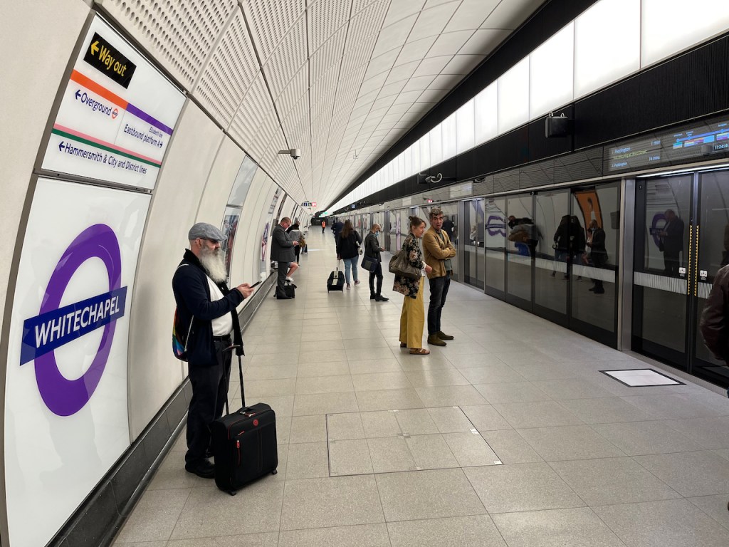 Passengers waiting on the very long and wide Elizabeth line platform at Whitechapel, with glass windows and doors along the platform edge.