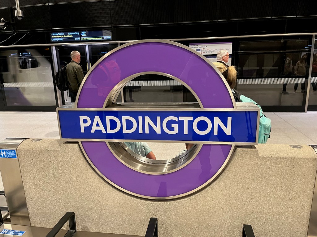 A large, thick, metal, purple roundel sign, consisting of a purple circle with a blue bar across it, for Paddington station. The bottom half of the purple circle is embedded into a wall between two benches.