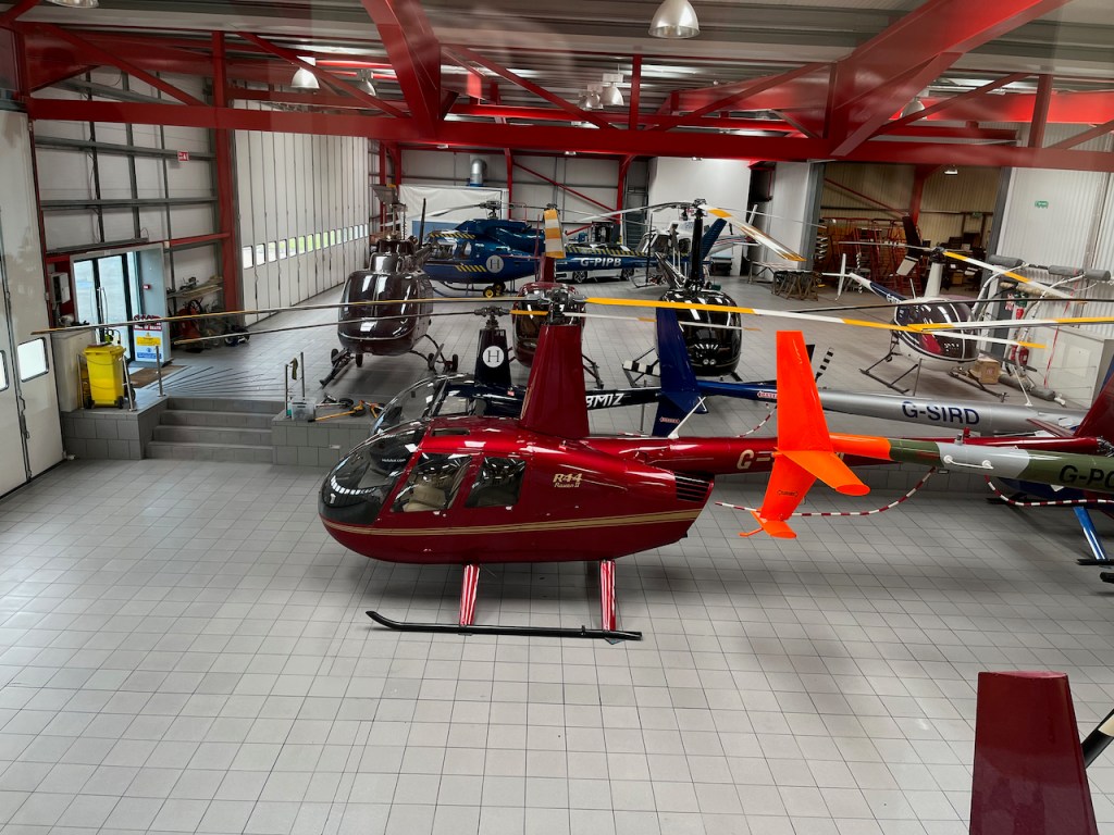 The hangar at Heli Air, containing helicopters with different colours of bodywork, including black and blue. The nearest helicopter to the camera is a red helicopter with a gold stripe along the side.