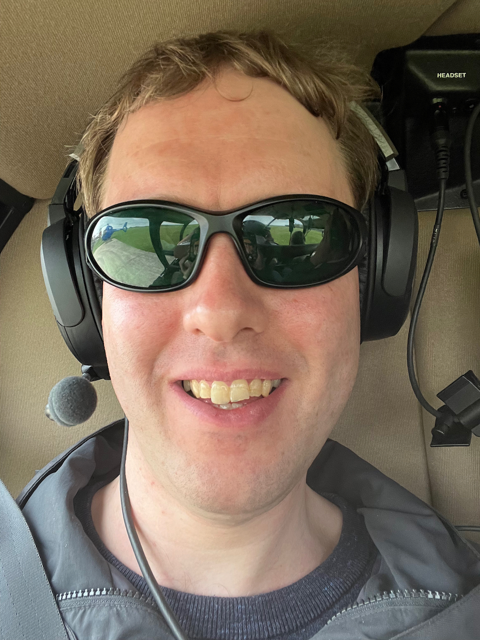 Selfie of Glen smiling while seated in the helicopter, wearing the headset that combines headphones and a microphone. His green sunglasses show a reflection of the front of the helicopter, and a blue helicopter parked nearby.