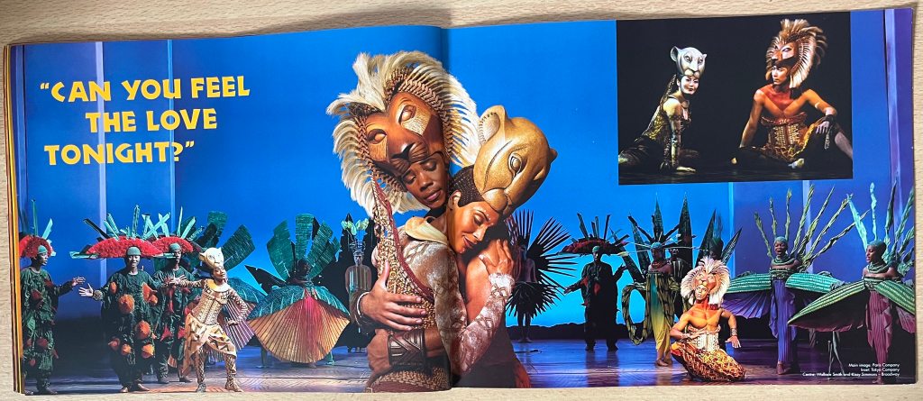 2-page spread from the programme for The Lion King, showing exquisitely dressed actors dancing on stage, while actors dressed as male and female lions hug each other in the centre. Text on the left page reads Can you feel the love tonight?