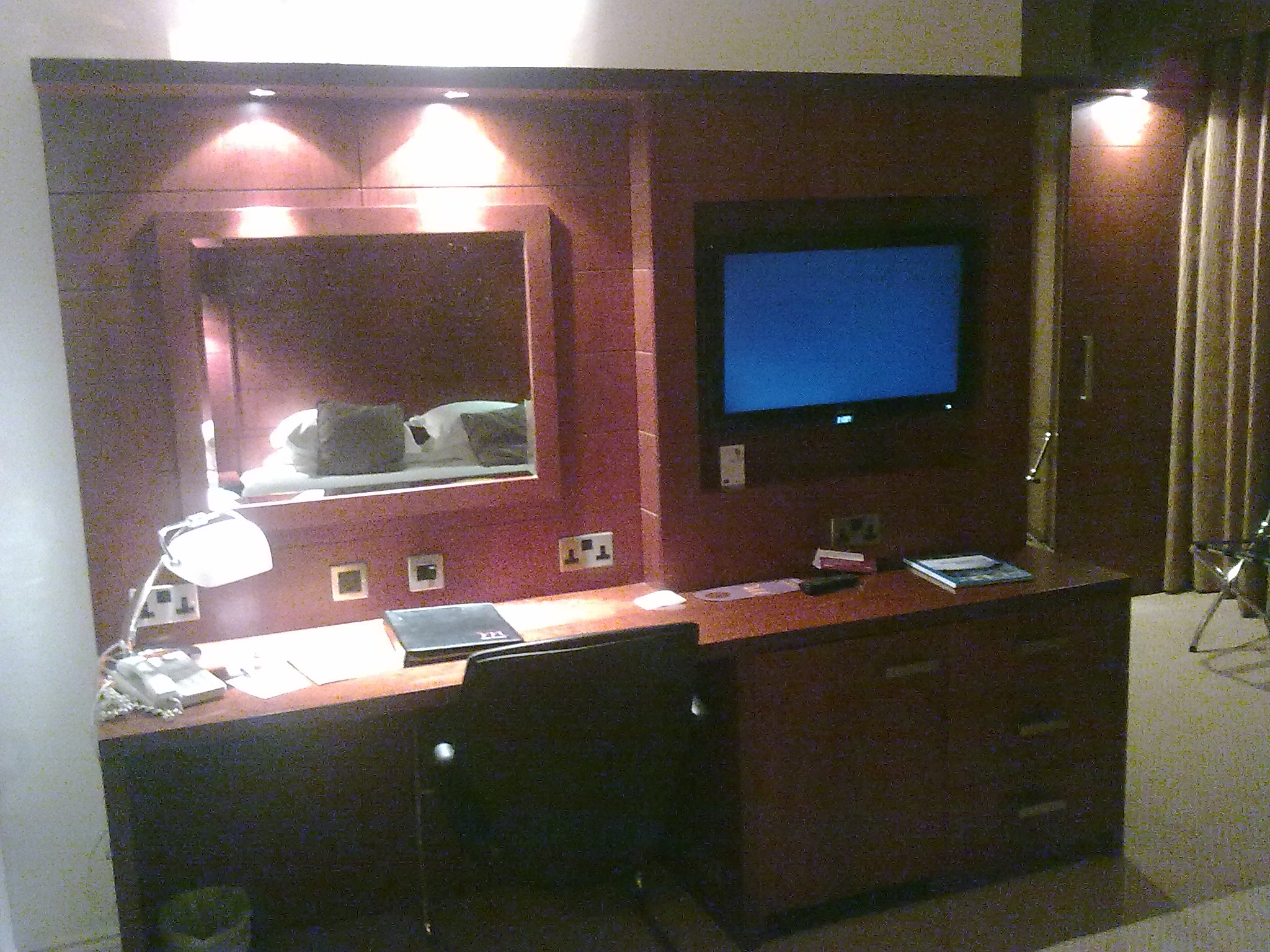 View of the desk in my Bristol hotel room, on which is a lamp, telephone and a folder of literature. On the wall above the desks are a mirror on the left, and a TV on the right, with plug sockets below them.