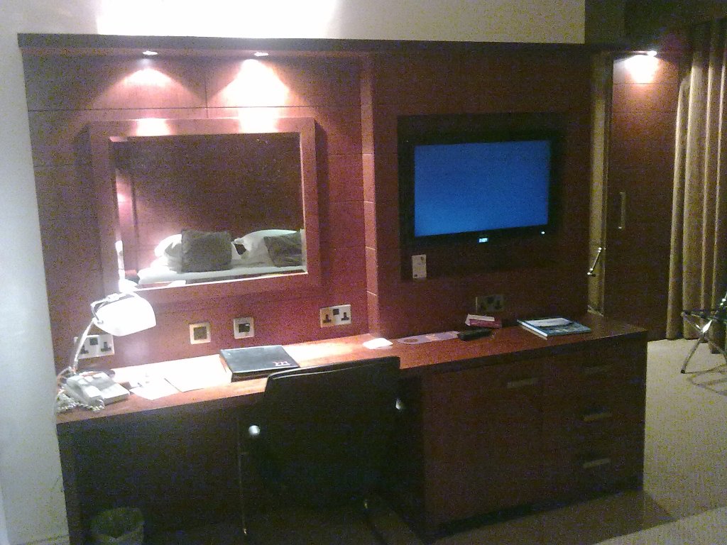 View of the desk in my Bristol hotel room, on which is a lamp, telephone and a folder of literature. On the wall above the desks are a mirror on the left, and a TV on the right, with plug sockets below them.