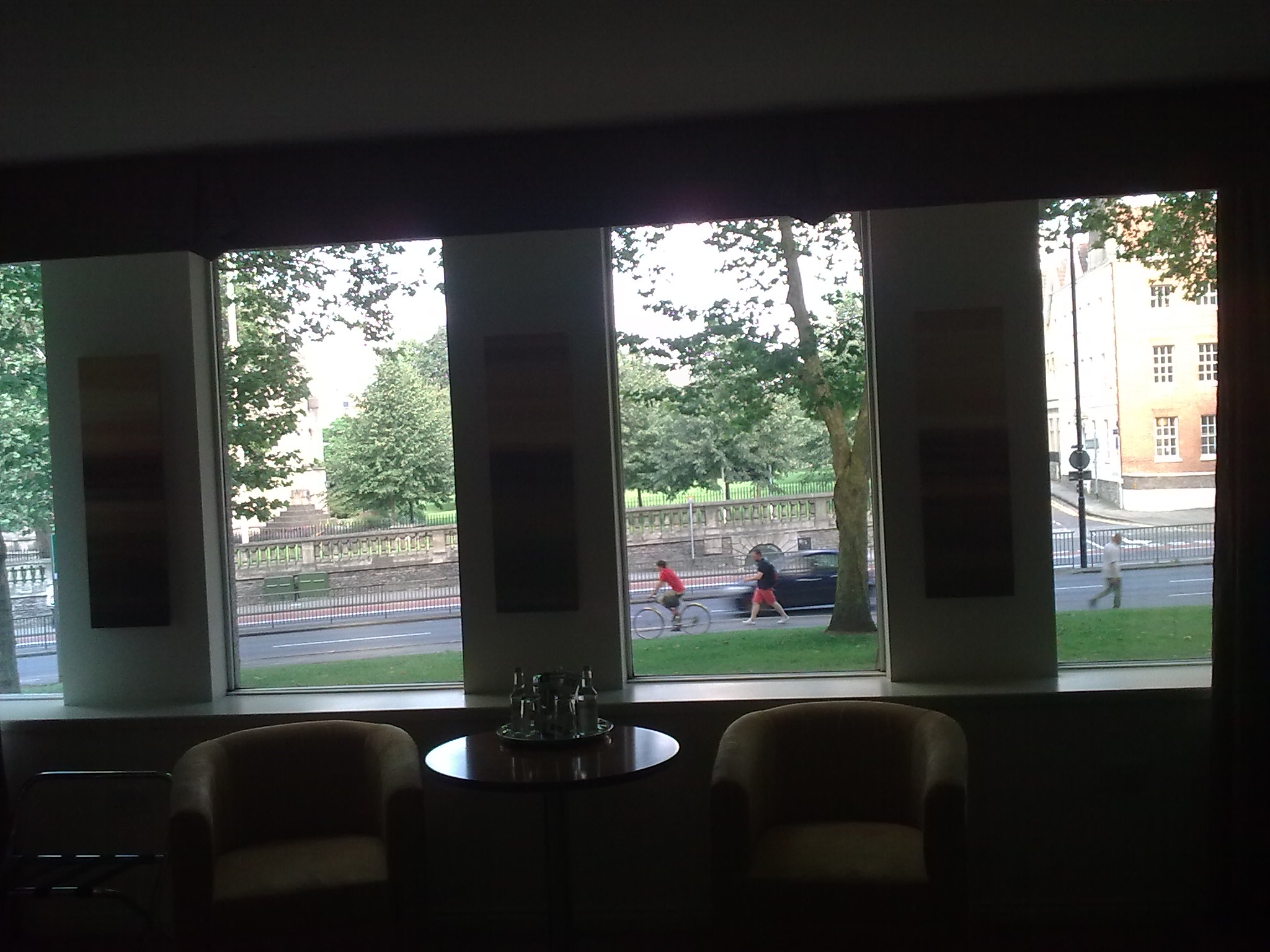 The view on to the street from my Bristol hotel room, with people cycling and walking outside. Across the road is a grassy area obscured by trees.