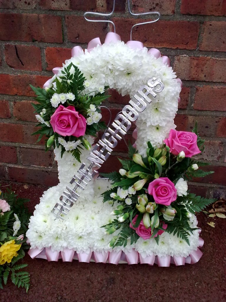 A large floral arrangement, with lots of white flowers forming an arch standing on a square base. Bunches of colourful pink and white flowers with green leaves are attached to the upper left and bottom right parts of the arch. In between them, stretching diagonally from bottom left up to top right, is silver capital lettering that says From The Neighbours.