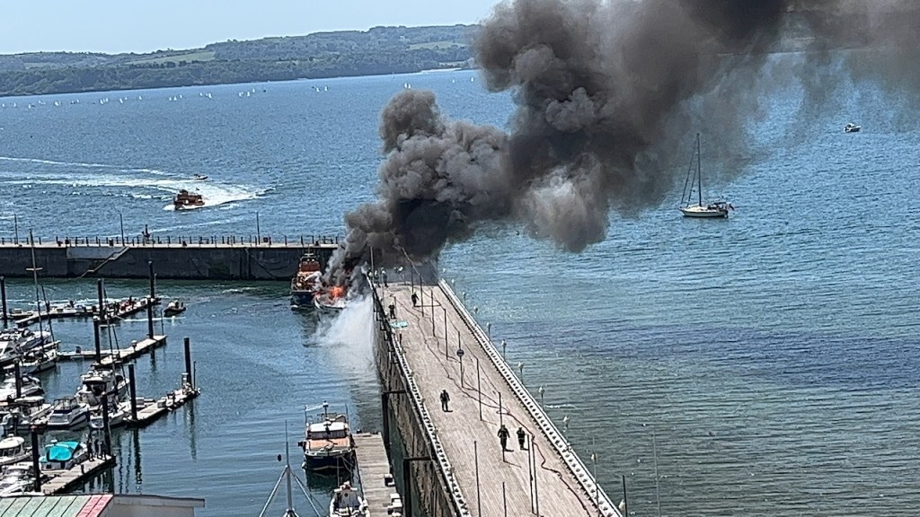 A super yacht on fire, billowing out a big cloud of black smoke, at the end of a long pier in Torquay harbour.