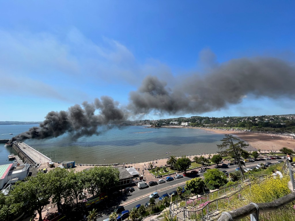 View of the huge cloud of black smoke from the burning super yacht, causing a long shadow on the sea and a beach in the distance as it travels into the distance.