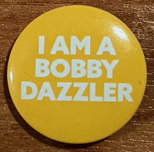 A small round yellow badge with white text that says I Am A Bobby Dazzler.