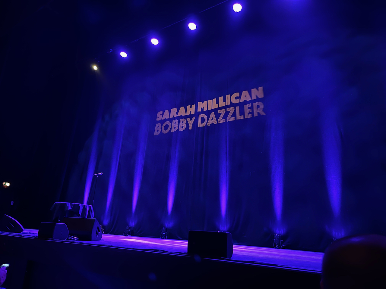 The large stage at the Hammersmith Apollo, all lit up in blue, with the title Sarah Millican Bobby Dazzler in big letters in the centre of the backdrop.