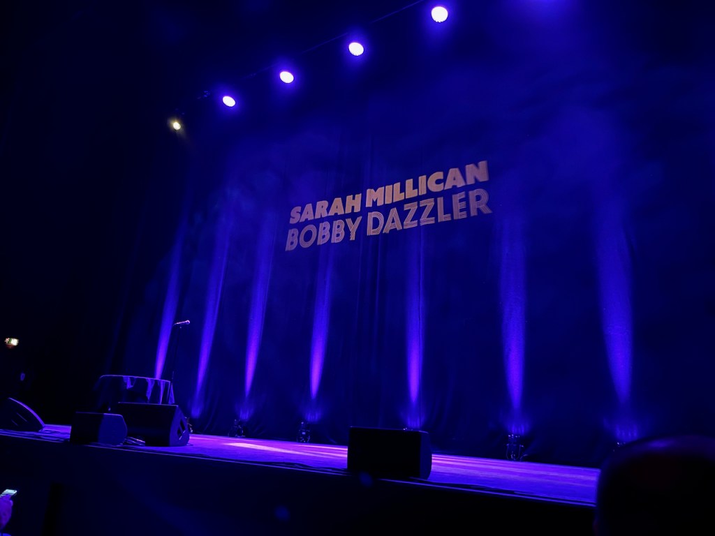 The large stage at the Hammersmith Apollo, all lit up in blue, with the title Sarah Millican Bobby Dazzler in big letters in the centre of the backdrop.