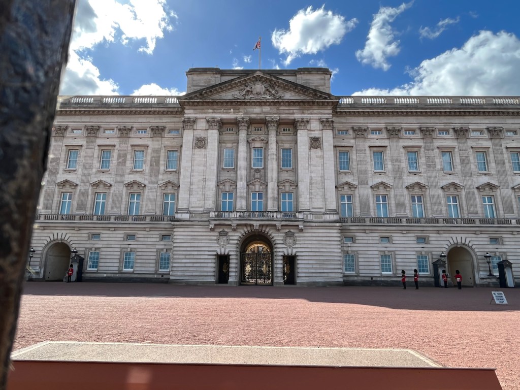 The central section of Buckingham Palace, showing the first floor balcony where the Royal Family sometimes appear. Below it is an archway shut off by an ornate gold gate.