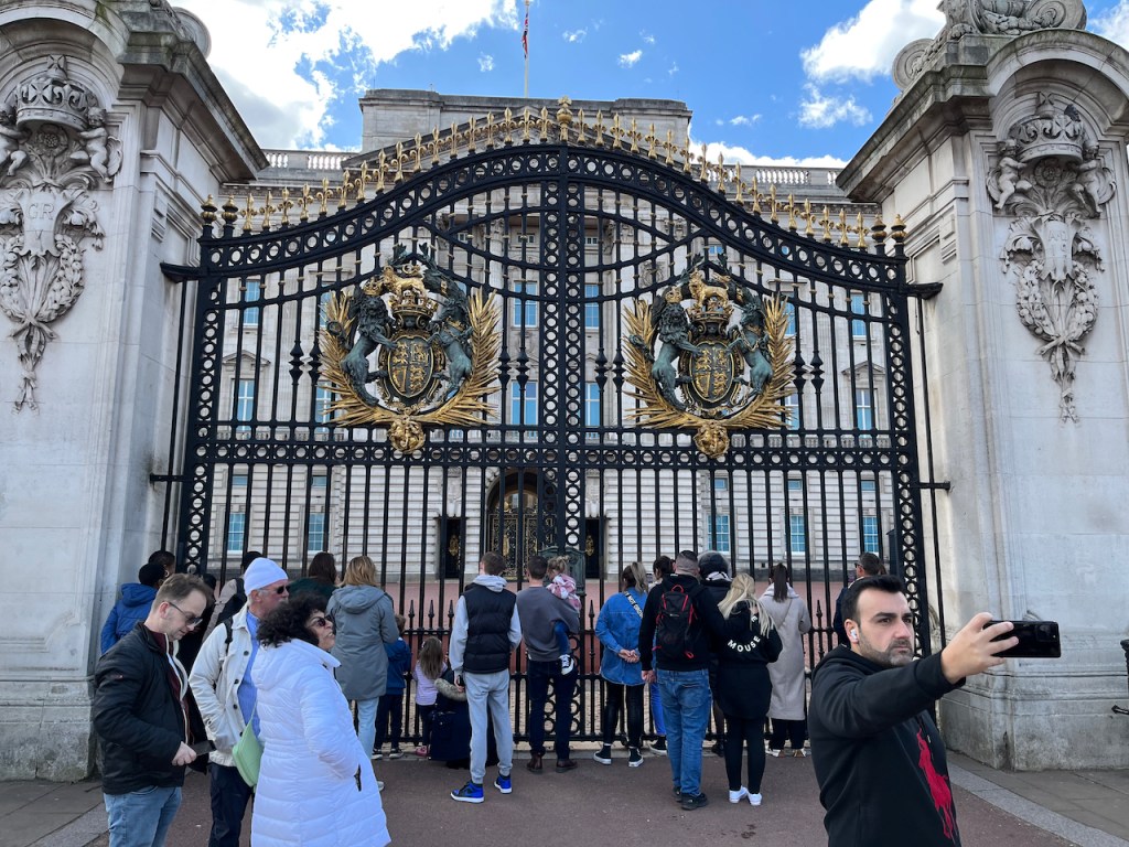 The large, black, ornate double gates to Buckingham Palace. The top of each gate, decorated with ornate gold spikes, curves up from each side so they meet as a curved peak in the centre. Lower down, each gate has a large ornate shield, with gold leaves around the sides, and a coat of arms in the centre flanked by a black lion wearing a gold crown on its left and a black horse on the right. A small gold lion sits on a gold crown on top of the coat of arms.