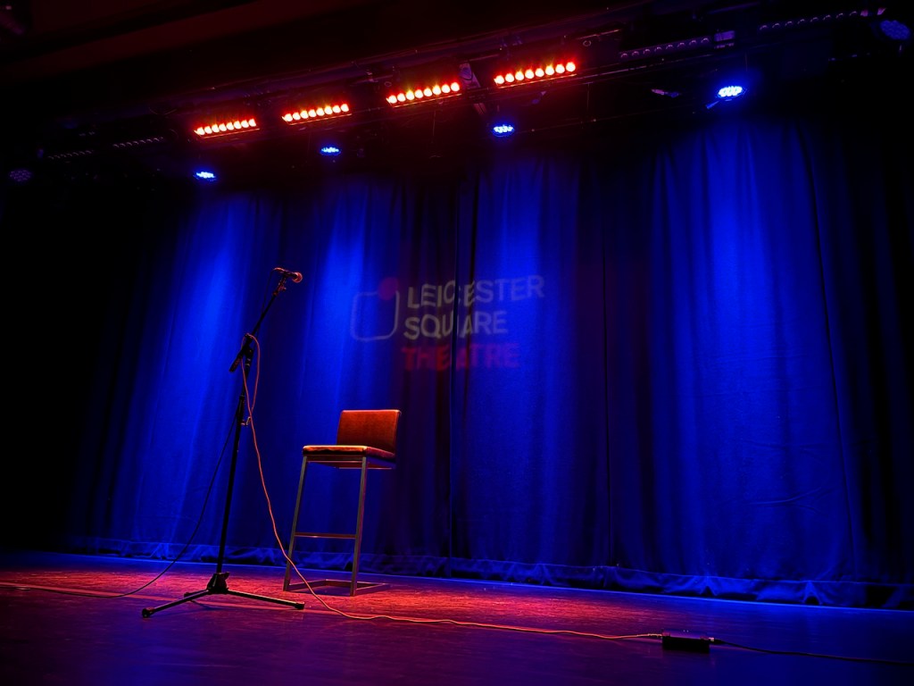 The stage of the Leicester Square Theatre, empty except for a tall chair with a low back, facing a tall microphone stand. The chair has just 2 legs at the front, connected to  square frames underneath the seat and on the floor that keep it upright. The backdrop is a curtain lit up in blue, with the words Leicester Square in white, and Theatre in red, projected upon it.