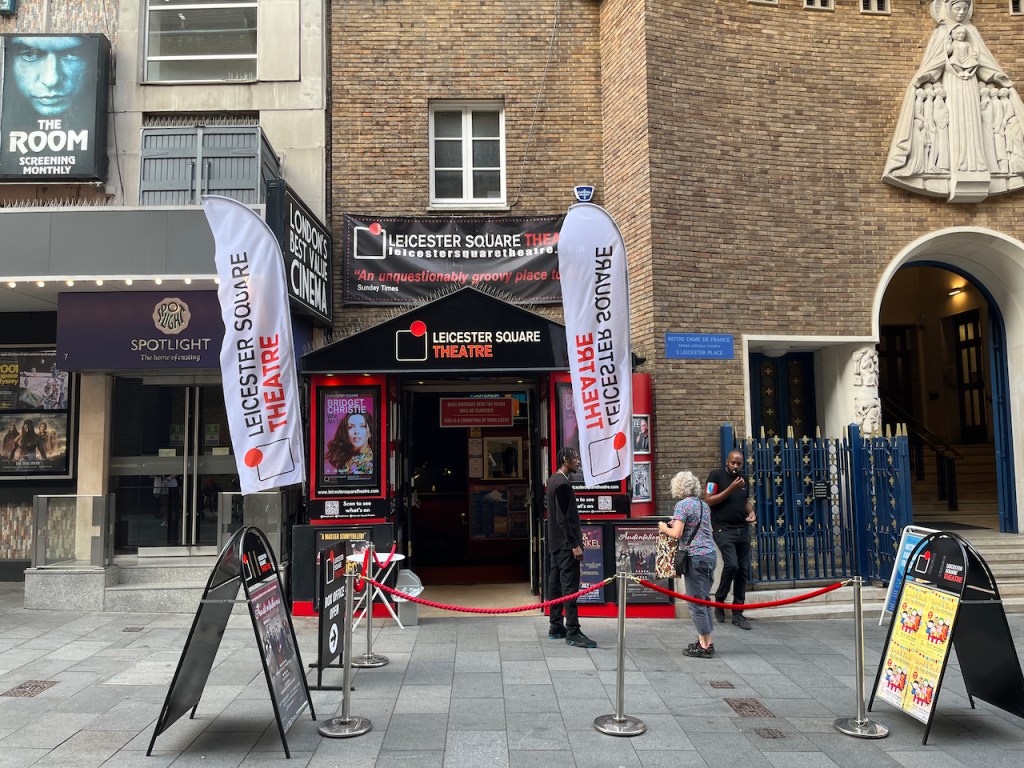 The entrance of the Leicester Square Theatre. A triangular black canopy above the entrance bears the theatre's name, as does a horizontal black banner above it, and 2 white vertical banners hanging down either side. The black banner above the entrance also includes their website address, and a quote from the Sunday Times that says it's an unquestionably groovy place.