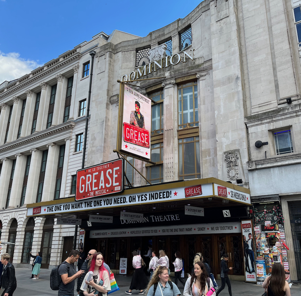 The outside of the Dominion Theatre, with signage for Grease The Musical. A rectangular red sign with white lettering above the entrance reads We Go Together, Grease The Musical, with Grease the largest word in the centre. Just below that, along the edge of the canopy overhanging the entrance in black lettering on white, text reads The One You Want, The One You Need, Oh Yes Indeed. A similar white strip directly over the doors of the theatre carries some of the best known song titles from the show.