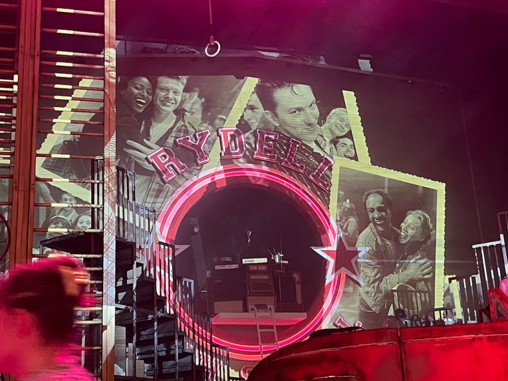 Close-up of the rear of the stage during the interval of Grease, In the centre of the circular Rydell High School logo, a large hole reveals the DJ booth with various items of equipment. Meanwhile, around the outside of the school logo are large black and white photos of people smiling and dancing together.
