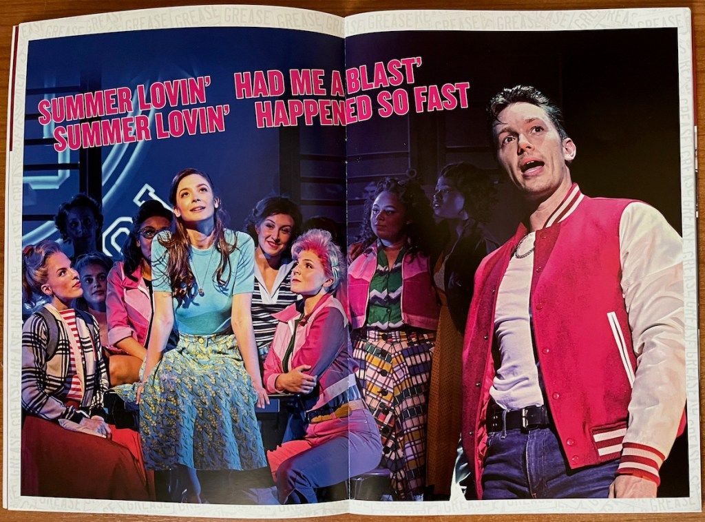 2-page spread from the programme for Grease The Musical. On the left page, Sandy's girl friends gather around her as she sits wearing a blue short sleeved top and flowery skirt, looking up thoughtfully. On the right page Danny stands alone singing, wearing a red shirt open over a white vest and blue jeans. Lyrics in pink above Sandy's photo reads Summer lovin', had me a blast, summer lovin', happened so fast.