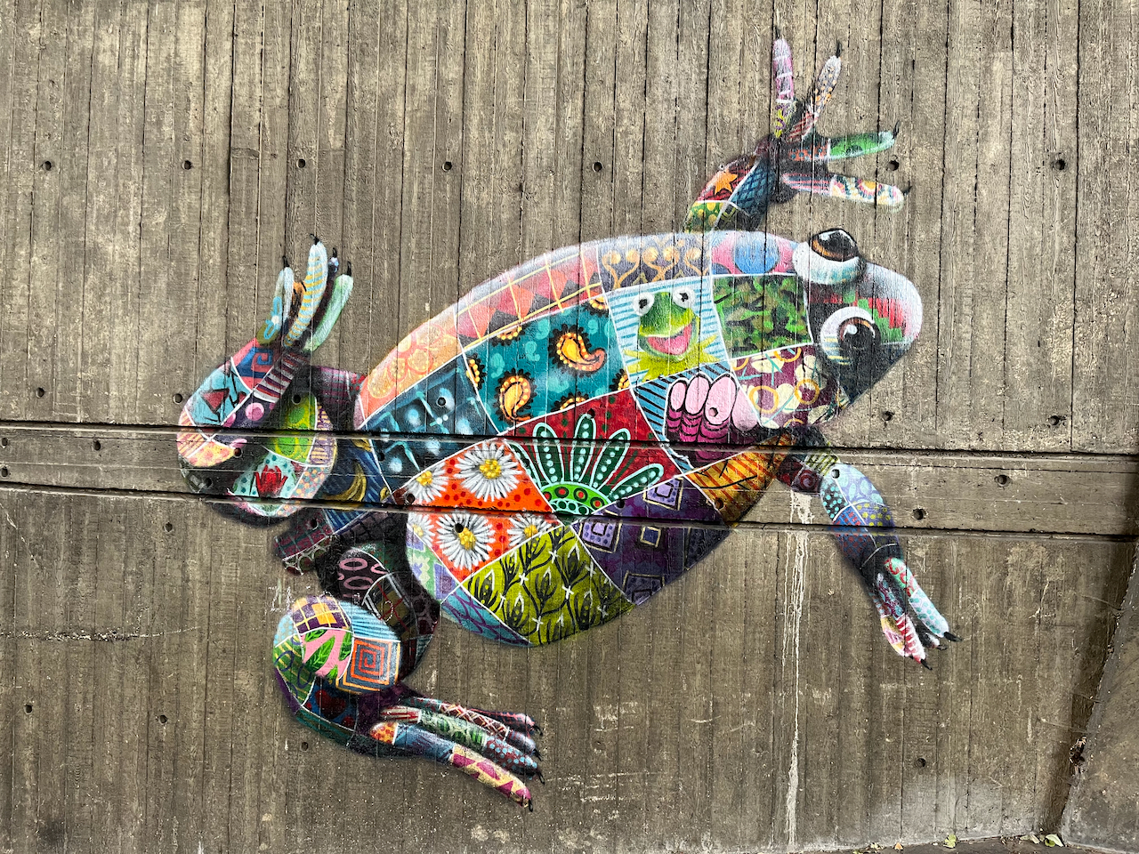 A mural of a Lake Oku Clawed Frog, made up of lots of different colourful patterned patches. One of the patches contains the smiling face of Kermit The Frog, while other patches contain flowers, leafy designs or other random patterns.