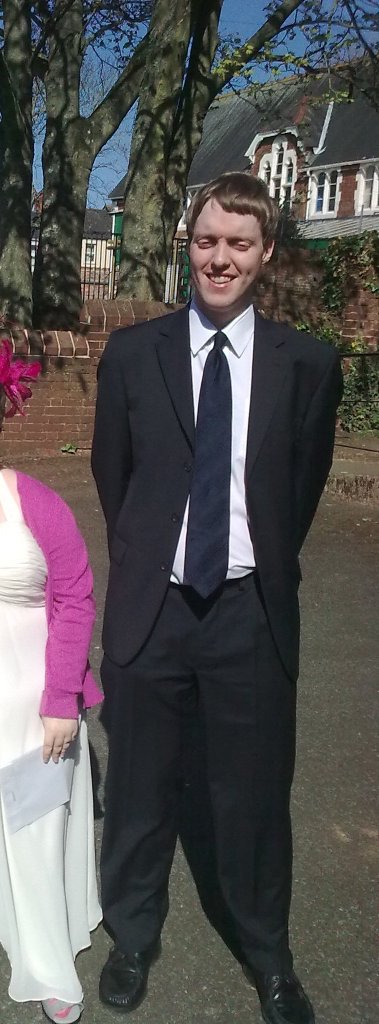Glen smiling and squinting in the sunlight, as he stands in a black suit with white shirt and navy tie.