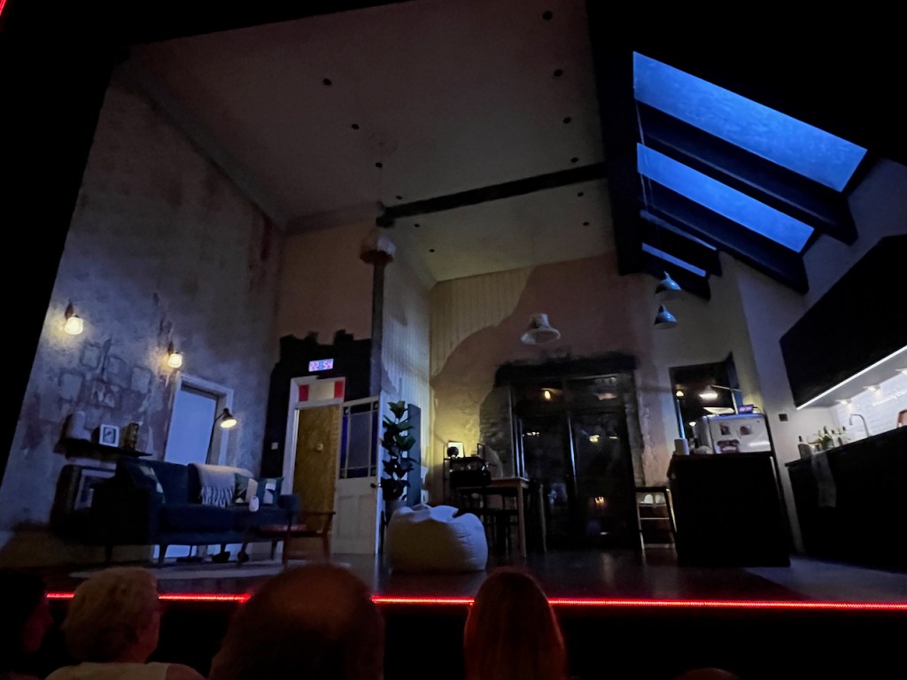 The stage for 2:22 A Ghost Story, showing an open plan living space. On the left is a living room, which looks dated and in need of renovation, while on the right is a new modern kitchen with skylights. In between, set further back, is a dining area in front of some patio doors. Also on the left, above an open doorway out of the living area, is a digital clock with red numbering, which shows the time throughout the show, as does a smaller portable clock on top of the fridge on the opposite side of the stage.