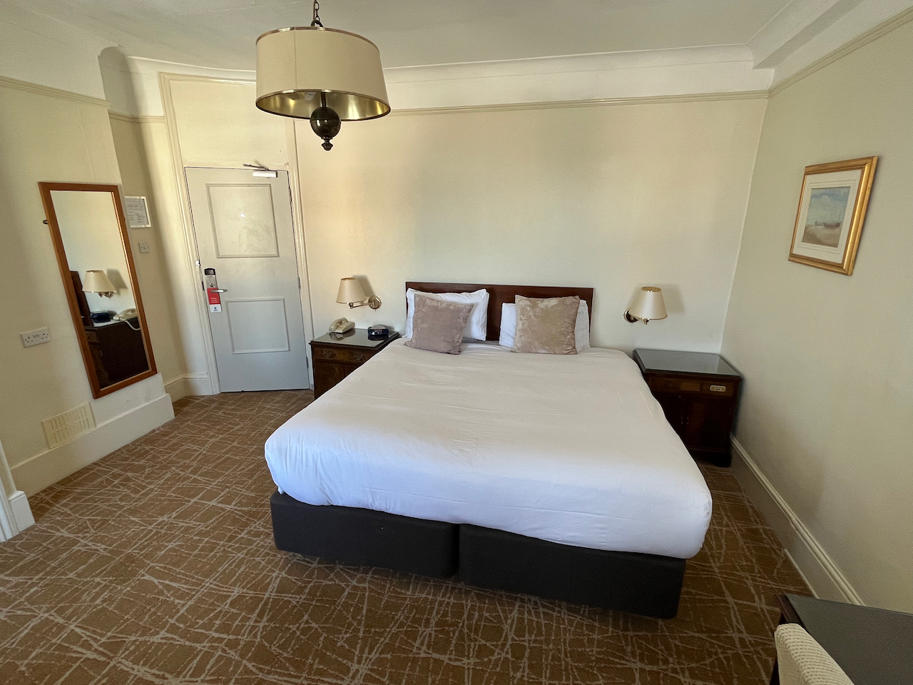 My bedroom in the Best Western Clifton hotel. The  double bed has dull coloured square cushions in front of the white pillows. Small wooden bedside cabinets sit on each side of the bed. A full-length mirror is on a wall to the left near the door.