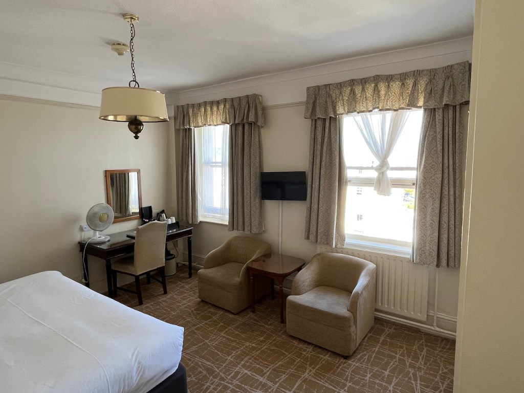 My bedroom in the Best Western Clifton hotel, looking towards the two large windows, the bottom half of the right window having been fully pulled up to let air in. A small TV screen is mounted on the wall in between the windows, and below it are 2 chairs either side of a wooden table. In the corner by the left window there is a desk and chair in front of a wall mirror. The desk has tea and coffee supplies, and a large fan.
