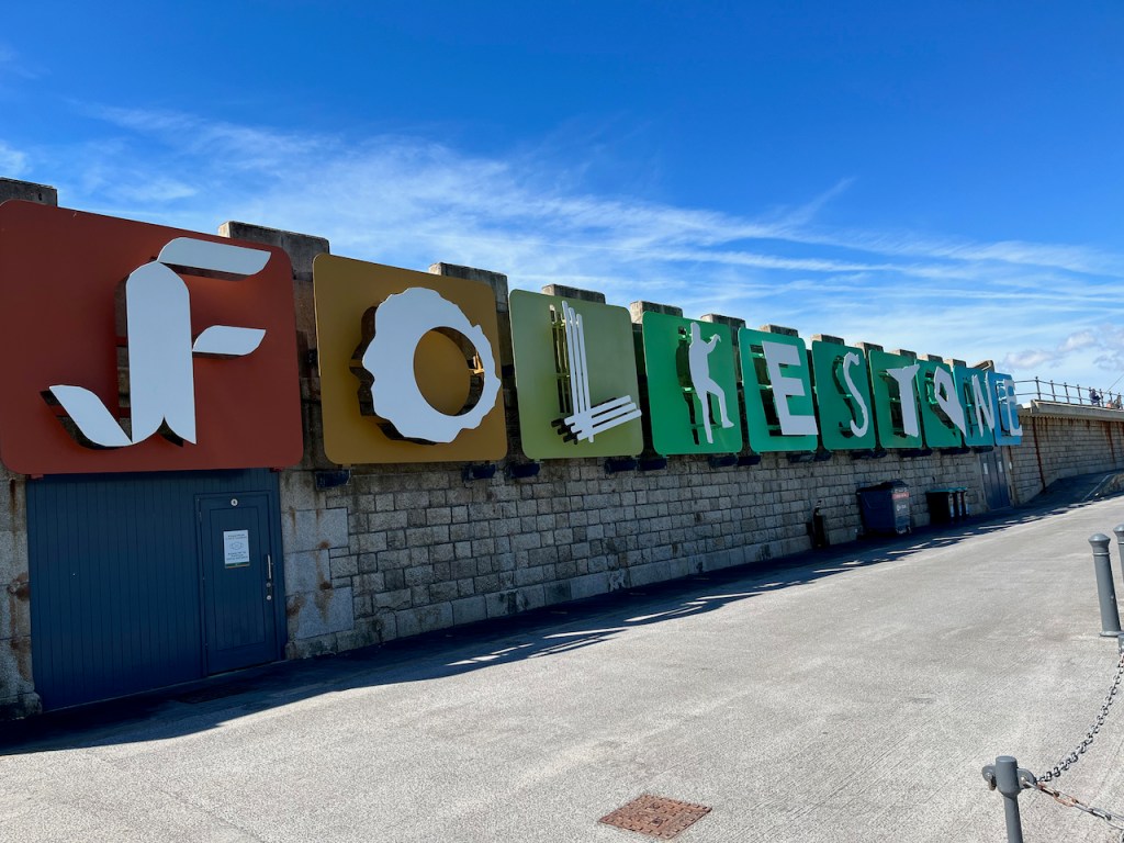 A long wall sign made up of very large coloured blocks that spell out the word Folkestone in stylised white letters. Each letter is styled differently, with the K in particular looking like a person holding up their left arm and leg to form the letter.