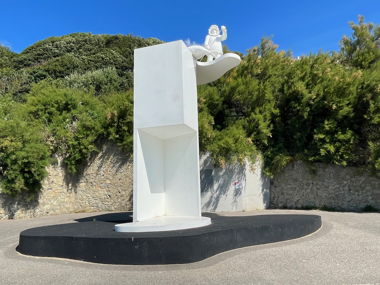 A sculpture of a tall white tower, with a ledge sticking out at the top, on which stands a white statue of an Inuit figure dressed in a thick wooly coat and hood, and holding a long stick. Next to them is a white statue of a seal laying on top of the tower so its tail hangs over the edge. The artwork is called The Ledge by Bill Woodrow.