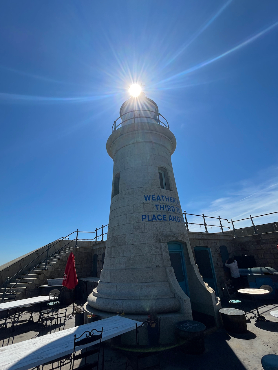 The lighthouse at the end of the Folkestone Harbour Arm, which is now a champagne bar, so there are tables and chairs around it. The photo is taken from such an angle that the sun is directly on top of the tower, making it look as if the lighthouse has its light on.