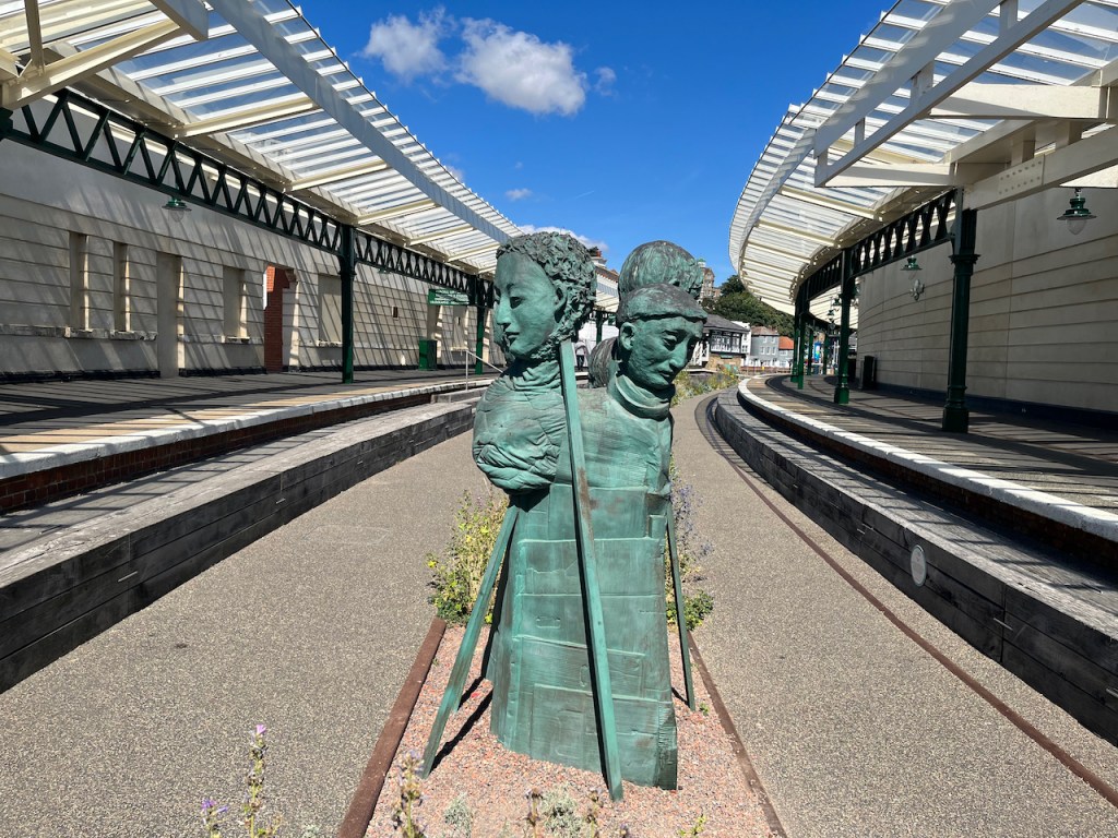 A sculpture of 4 people standing together in the middle of the former railway bed, which is now open to pedestrians to walk along, at the old Folkestone Harbour Station. The artwork is called Rug People by Paloma Varga Weisz.