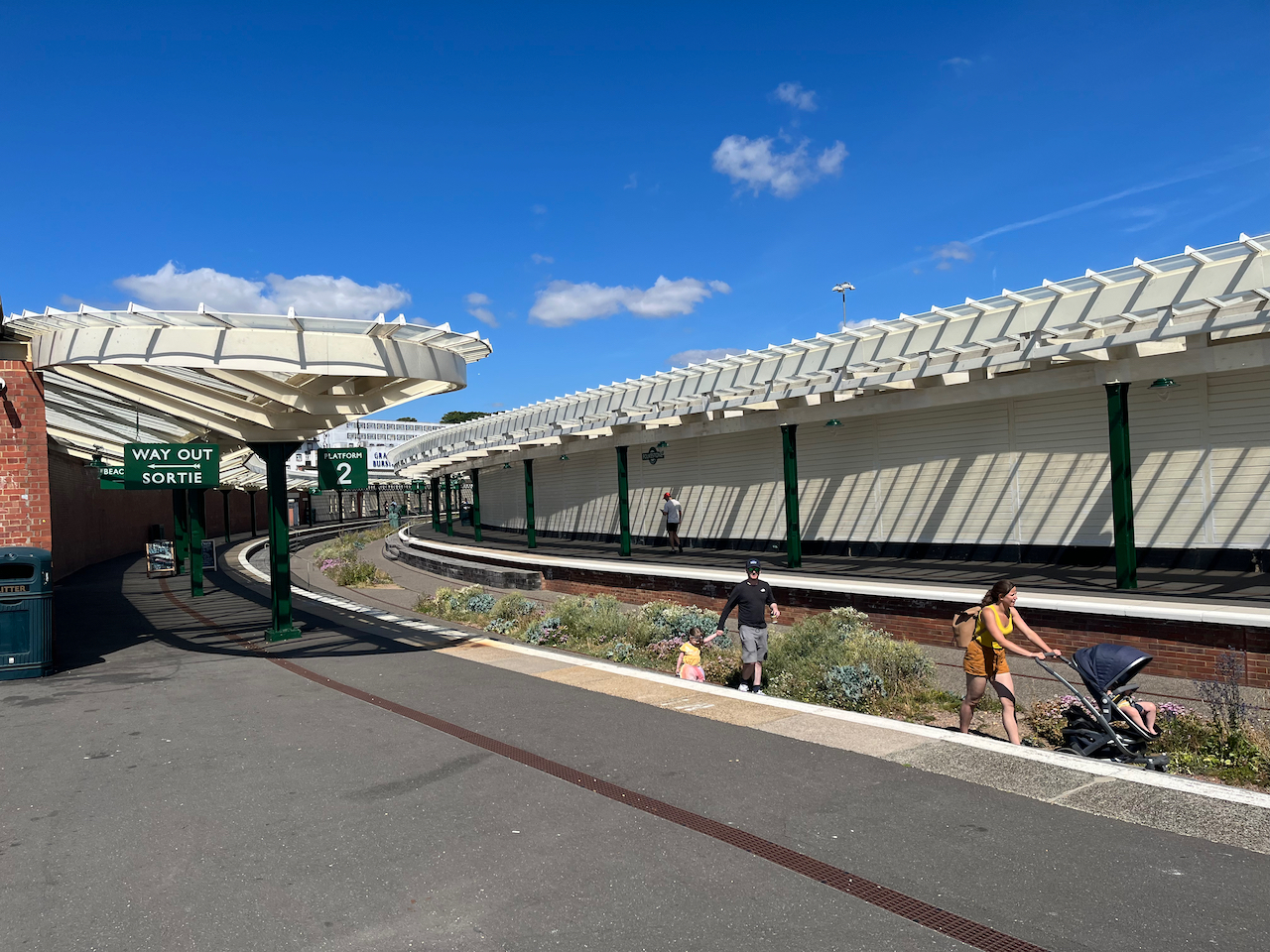 Folkestone Harbour Station with its curving platforms, and signage in both English and French. The railway itself has been removed, and replaced with footpaths either side of greenery planted down the centre, so you can see people walking along and pushing prams.