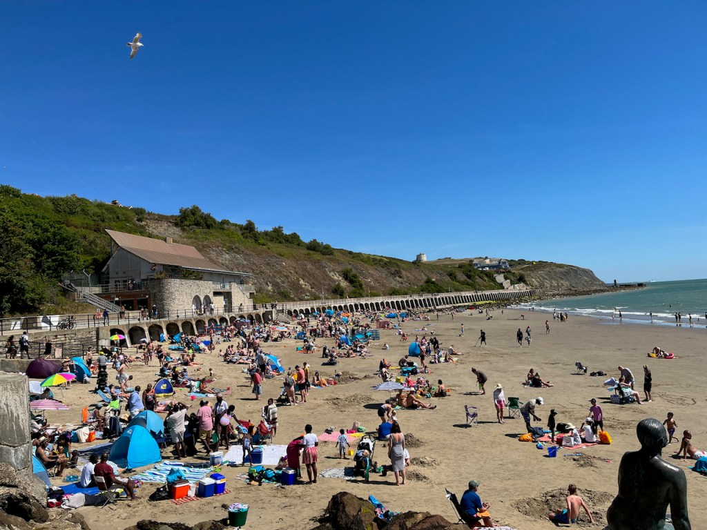 Sunny Sands beach in Folkestone, beneath a clear blue sky, full of families and other people enjoying the sunshine.