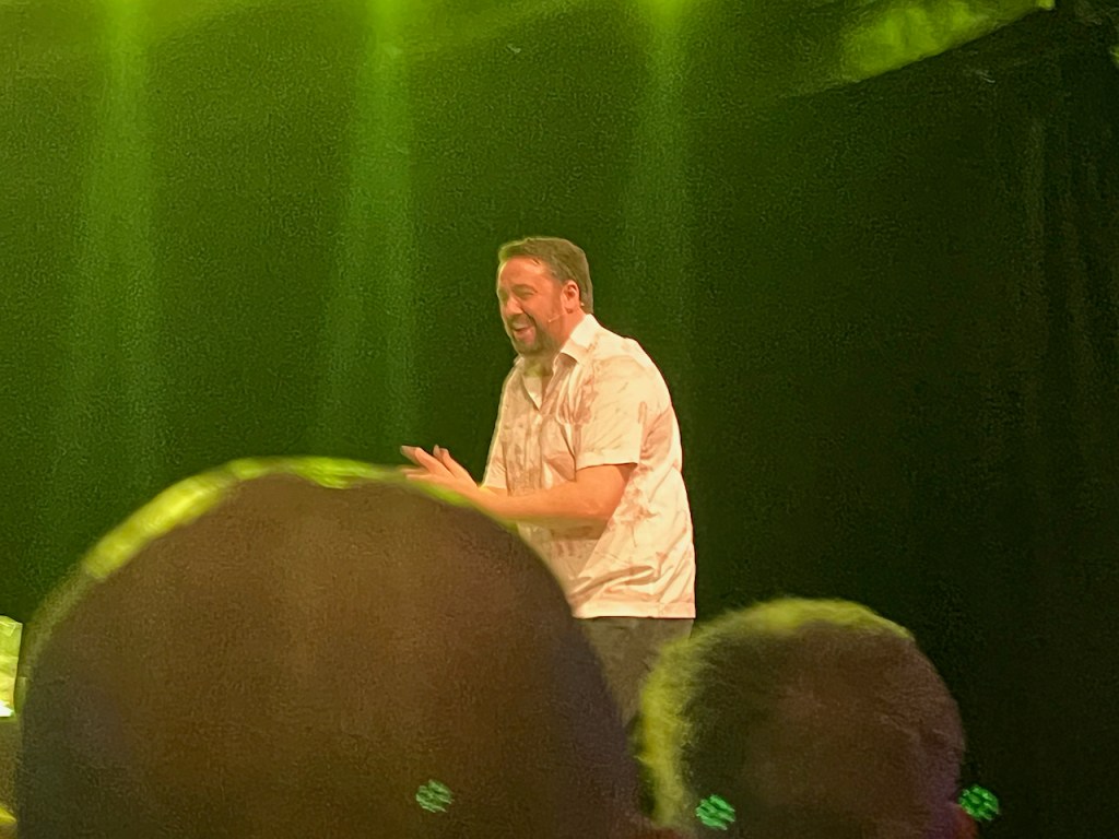 Jason Manford smiling as he performs on stage in a white short sleeved shirt and dark trousers.
