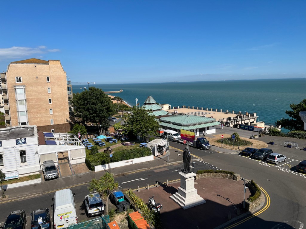 The Leas Cliff Hall theatre, a building with a flat roof on the right end and a large pyramid shaped roof on the left. A large terrace behind it, overlooking the sea, has a large chessboard design in the middle of it, viewed from upstairs in a nearby hotel.